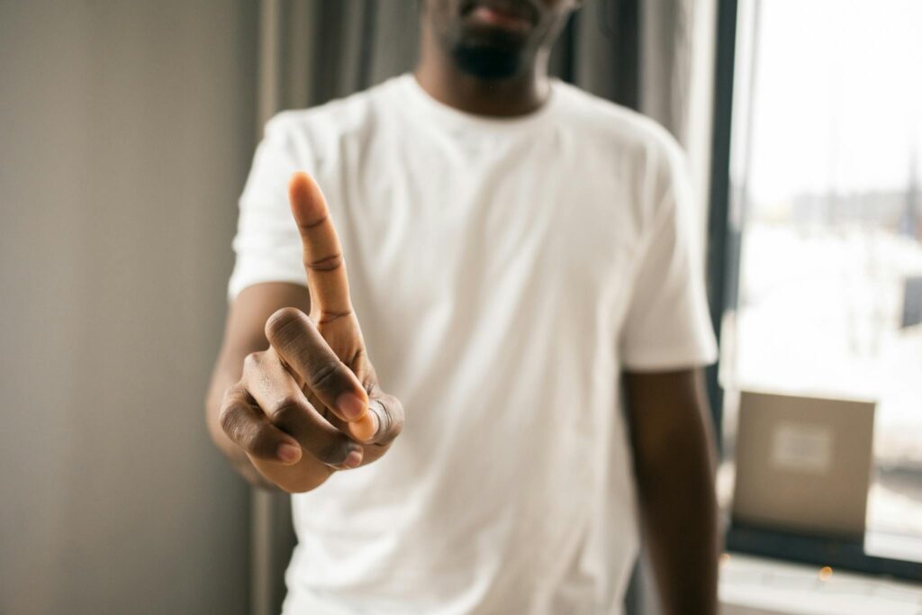 Crop anonymous African American male in white t shirt showing stop gesture with index finger pointing up in light room on blurred background