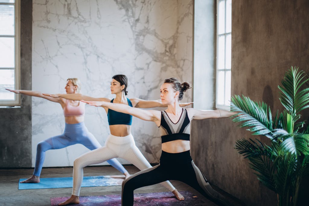 Three women practicing Warrior II yoga pose indoors, promoting fitness and mindfulness.